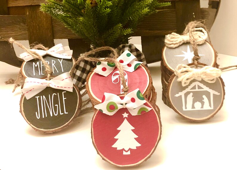 May include: Set of six wood slice Christmas ornaments with various designs. The ornaments are painted in red, gray, and black with white designs. The designs include a Christmas tree, a candy cane, a star, a nativity scene, and the words "Merry Jingle".