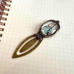Puede incluir: Marcapáginas de estilo vintage con imagen ovalada de un conejo con chaqueta azul. El marcapáginas tiene un acabado de latón antiguo y descansa sobre una página de cuaderno cuadriculada. La ilustración del conejo recuerda a un libro infantil clásico.