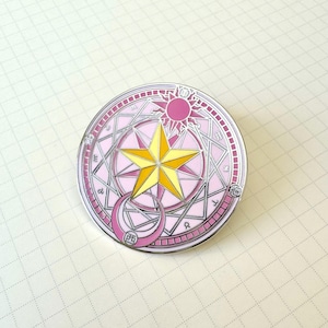 Puede incluir: Un pin redondo de esmalte rosa y plateado con una estrella amarilla en el centro. El diseño incluye una luna creciente, un sol y patrones geométricos. El pin está sobre un fondo de papel blanco rayado.