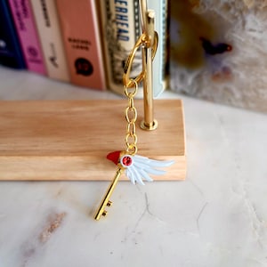 May include: Gold key charm with a red and white wing design. The key has a red gem on the top and a white wing on each side. The charm is attached to a gold chain and a gold key ring.