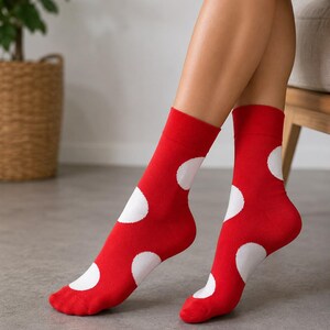 Könnte beinhalten: Ein Paar wadenhohe Socken in leuchtendem Rot mit großen, kreisförmigen weißen Punkten, die über den Stoff verteilt sind. Die Socken haben ein geripptes Bündchen am oberen Rand und bedecken die Füße und Unterschenkel mit einem gleichmäßigen Muster.
