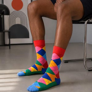May include: A pair of crew-length socks featuring a vibrant argyle pattern in shades of red, yellow, pink, blue, and green. The socks have a solid red cuff at the top and a solid light blue toe section, with thin black lines forming the diamond shapes.