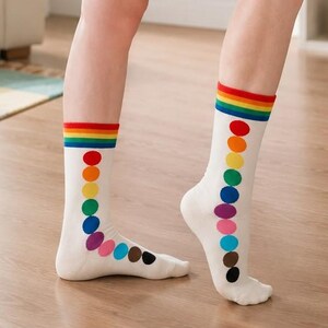 Könnte beinhalten: Ein Paar weiße Socken mit einem horizontalen Regenbogenstreifen am oberen Bündchen. Unter dem Streifen befindet sich eine vertikale Reihe aus bunten, kreisförmigen Punkten in Rot, Orange, Gelb, Grün, Blau, Violett, Pink, Hellblau, Braun und Schwarz.