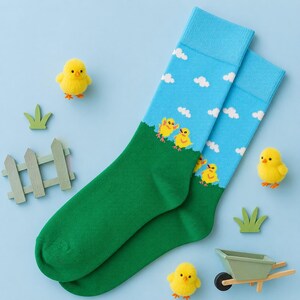Puede incluir: Un par de calcetines con un cielo azul con nubes blancas y un campo de hierba verde con pollitos amarillos. Los calcetines están rodeados de pequeñas figuras de pollitos amarillos, piezas de valla de madera verde y una carretilla verde.