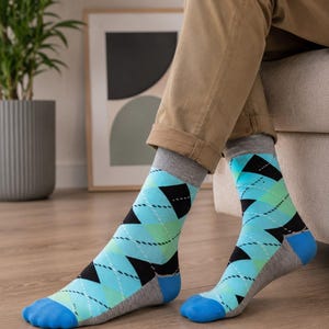 May include: A pair of crew-length socks featuring a repeating argyle pattern in shades of light blue, green, and black. The socks have a grey cuff, a solid blue toe section, and a grey heel, accented with thin white dashed lines across the diamonds.