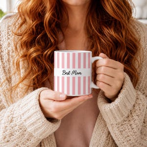 Op de afbeelding: Een witte keramische mok met verticale lichtroze strepen en een effen witte horizontale band in het midden. De tekst "Best Mom" is in een zwart cursief lettertype op de witte band gedrukt. De mok heeft een gebogen handvat aan de rechterkant.