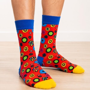 Könnte beinhalten: Ein Paar Socken mit einem leuchtend roten Grund und einem sich wiederholenden Muster aus bunten Blumen in Gelb, Blau, Grün und Pink. Die Socken haben einfarbige blaue Bündchen am oberen Rand und einfarbige gelbe Zehenbereiche.