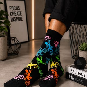 Könnte beinhalten: Ein Paar schwarze Socken mit einem sich wiederholenden Muster aus bunten Geckos in den Farben Pink, Blau, Grün, Gelb und Orange. Die Geckos sind in einem flachen, grafischen Stil mit geringelten Schwänzen über die gesamte Socke verteilt dargestellt.