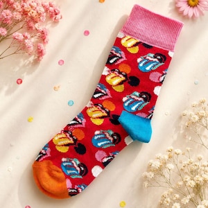 Könnte beinhalten: Ein roter Socken mit einem wiederkehrenden Muster aus stilisierten Mund- und Zungenlogos in Gelb, Blau und Weiß. Der Socken hat ein geripptes rosa Bündchen, eine leuchtend blaue Ferse und eine einfarbig orangefarbene Spitze für einen bunten Look.