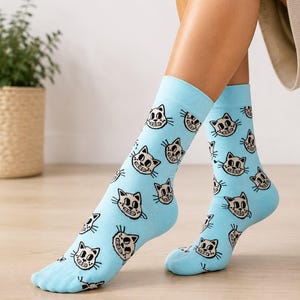 Könnte beinhalten: Hellblaue Socken mit einem sich wiederholenden Muster aus kleinen, lächelnden Katzengesichtern mit schwarzen Umrissen und cremefarbenen Fellakzenten. Die Socken haben ein geripptes Bündchen am oberen Rand und bedecken die Füße sowie die Unterschenkel.