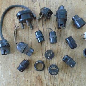 May include: A selection of antique electrical parts, including black lamp holders, plugs, and sockets, displayed on a wooden surface. The components are of various sizes and shapes, some with screw fittings and others with exposed wiring.