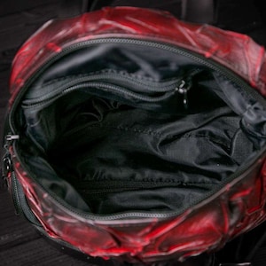 May include: Red leather backpack with a black zipper and a black lining. The backpack has a large main compartment and a smaller pocket on the front.