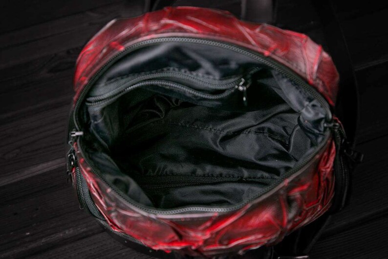 May include: Red leather backpack with a black zipper and a black lining. The backpack has a large main compartment and a smaller pocket on the front.