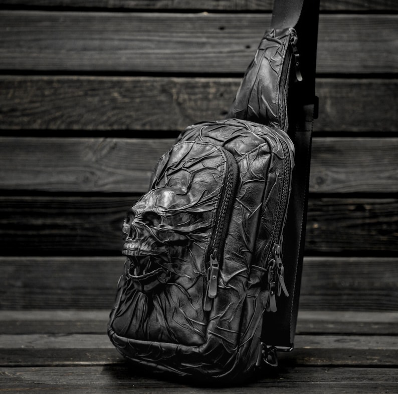 May include: A black leather crossbody bag with a skull design. The bag has two zippered compartments and a long adjustable strap.