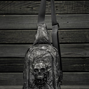 May include: Black leather crossbody bag with a skull design. The bag has a zipper closure and an adjustable strap.