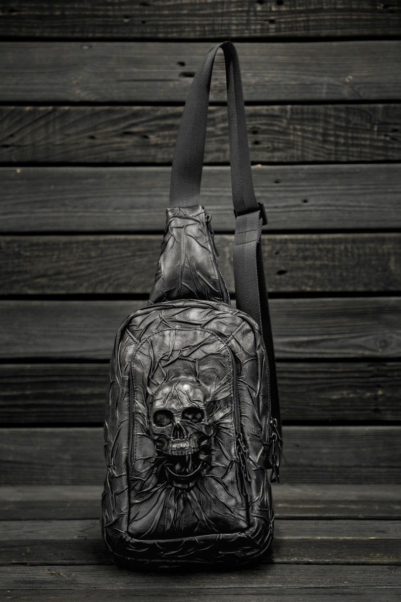May include: Black leather crossbody bag with a skull design. The bag has a zipper closure and an adjustable strap.