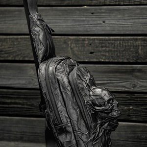 May include: A black leather backpack with a skull design. The backpack has a single strap and a zippered pocket on the front.