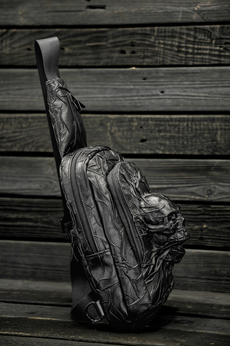 May include: A black leather backpack with a skull design. The backpack has a single strap and a zippered pocket on the front.