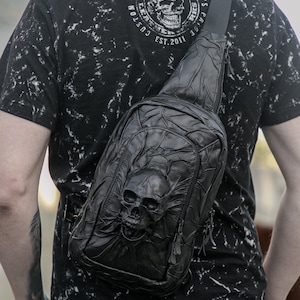 May include: A black leather crossbody bag with a skull design. The bag has a zipper closure and an adjustable strap. The bag is worn by a person wearing a black and white t-shirt with the text "Custom Leather for Bikers Genk Leather Works Est. 2011".