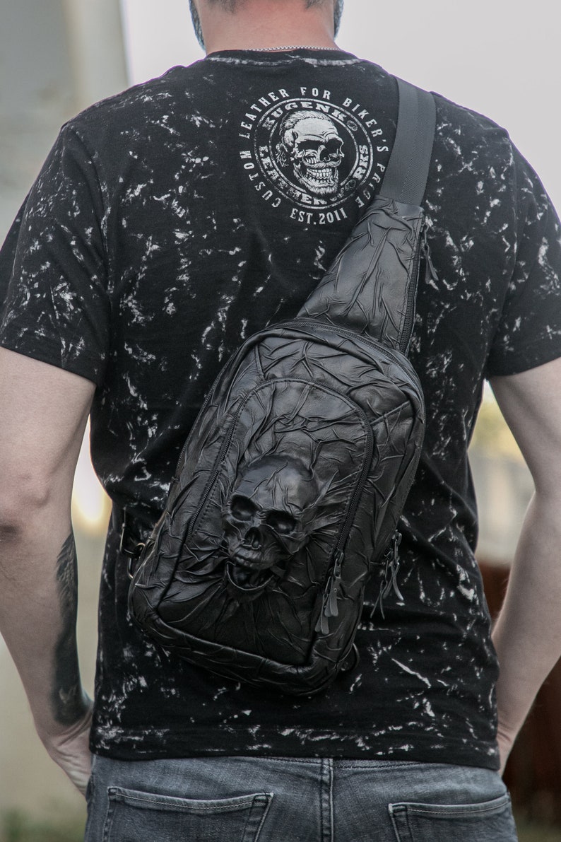 May include: A black leather crossbody bag with a skull design. The bag has a zipper closure and an adjustable strap. The bag is worn by a person wearing a black and white t-shirt with the text "Custom Leather for Bikers Genk Leather Works Est. 2011".