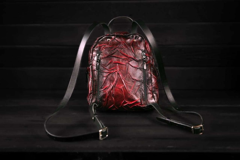 May include: A red leather backpack with black straps and buckles. The backpack has a textured, wrinkled design.