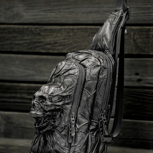 May include: Black leather crossbody bag with a skull design. The bag has two zippered compartments and a long adjustable strap.