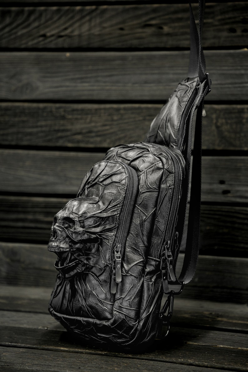May include: Black leather crossbody bag with a skull design. The bag has two zippered compartments and a long adjustable strap.