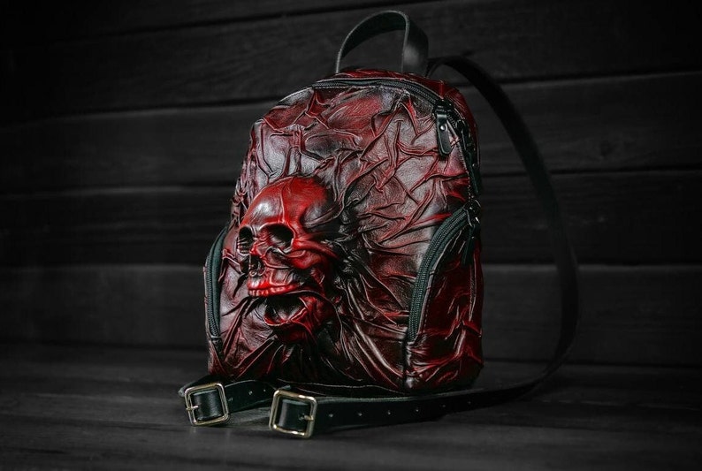 May include: A red leather backpack with a skull design. The backpack has a black strap and a zipper closure.