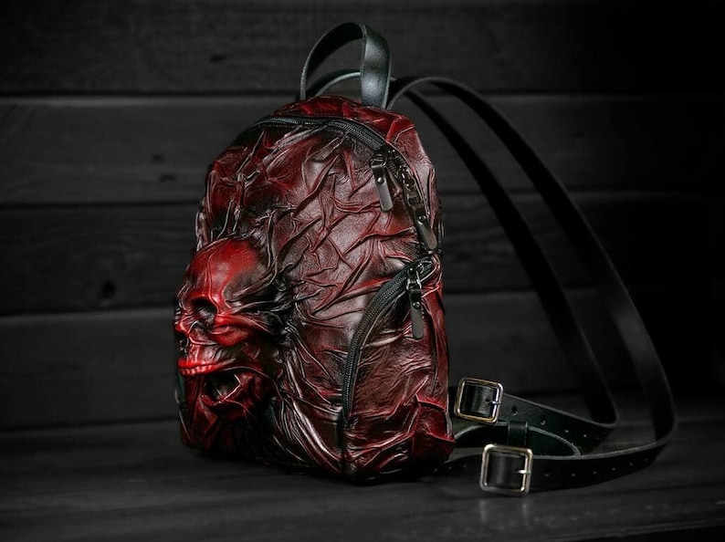 May include: A red leather backpack with a skull design. The backpack has black straps and buckles.