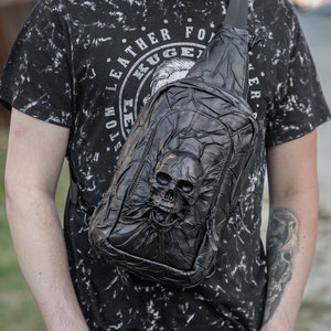 May include: A black leather crossbody bag with a skull design. The bag has a zipper closure and an adjustable strap. The bag is worn by a person wearing a black t-shirt with white print.