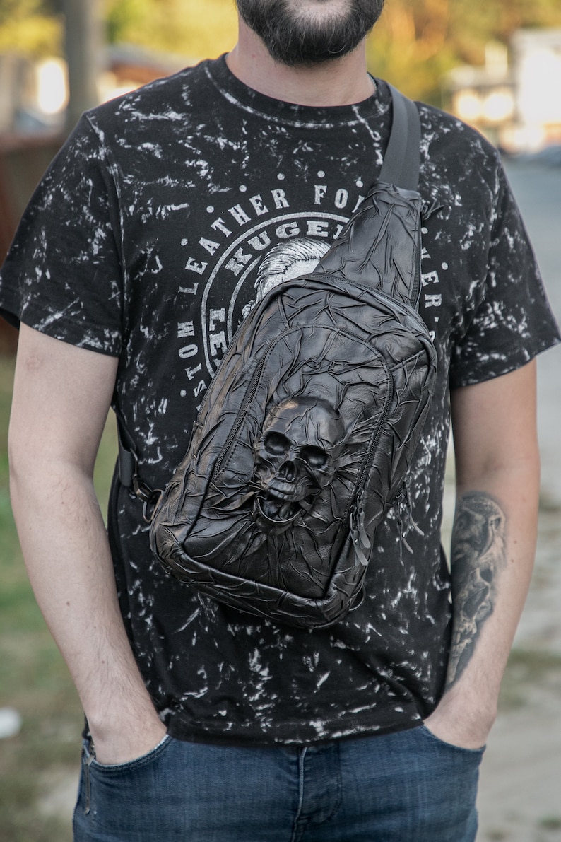 May include: A black leather crossbody bag with a skull design. The bag has a zipper closure and an adjustable strap. The bag is worn by a person wearing a black t-shirt with white print.
