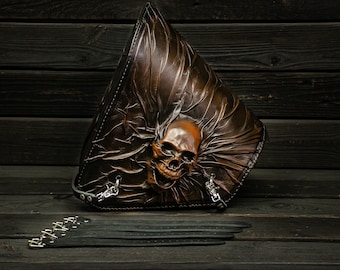 May include: A brown leather motorcycle saddlebag with a skull design. The bag has a flap with a silver buckle and a chain strap.