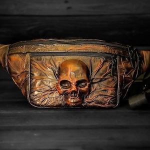 May include: A brown leather fanny pack with a skull design. The skull is embossed on the front of the bag and has a detailed design.