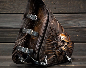 May include: A brown leather motorcycle saddlebag with a skull design. The bag has multiple buckles and straps.
