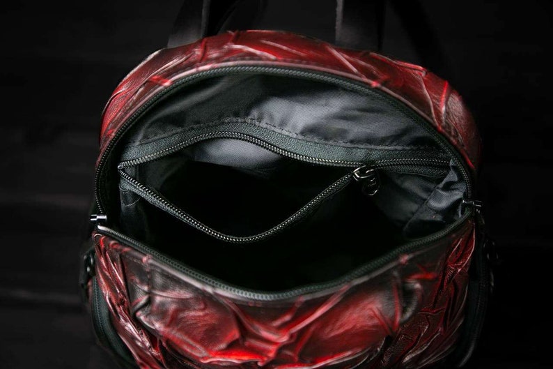 May include: Red leather backpack with a zippered pocket and a black lining. The backpack is open and the inside is visible.