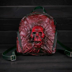 May include: A red leather backpack with a skull design. The skull is embossed on the front of the bag and is painted red. The backpack has green straps and buckles.