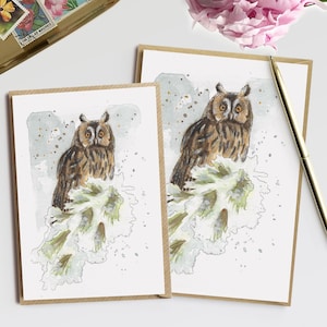May include: Two greeting cards with watercolor illustrations of owls perched on a branch. The cards have a brown border. A gold pen and a pink peony are next to the cards. A gold tray with vintage stamps is also in the image.