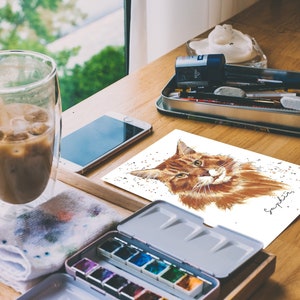 May include: A watercolor painting of a brown tabby cat with green eyes. The cat is looking directly at the viewer. The painting is on a white background with a few specks of gold paint. The artist's signature, "Sapphire", is in the bottom right corner.