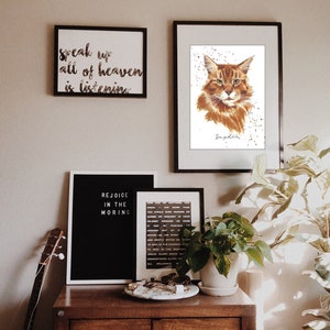 May include: A brown wooden dresser with three drawers, a black framed sign that says "Rejoice in the Morning", a white framed sign that says "Speak up all of heaven is listening", a black framed print of a cat with the name "Saphira" written below, a plant in a white pot, and a guitar leaning against the dresser.