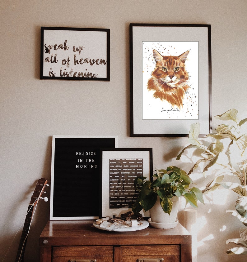 May include: A brown wooden dresser with three drawers, a black framed sign that says "Rejoice in the Morning", a white framed sign that says "Speak up all of heaven is listening", a black framed print of a cat with the name "Saphira" written below, a plant in a white pot, and a guitar leaning against the dresser.