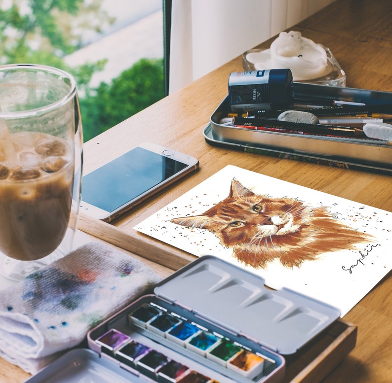 May include: A watercolor painting of a brown tabby cat with green eyes. The cat is looking directly at the viewer. The painting is on a white background with a few specks of gold paint. The artist's signature, "Sapphire", is in the bottom right corner.