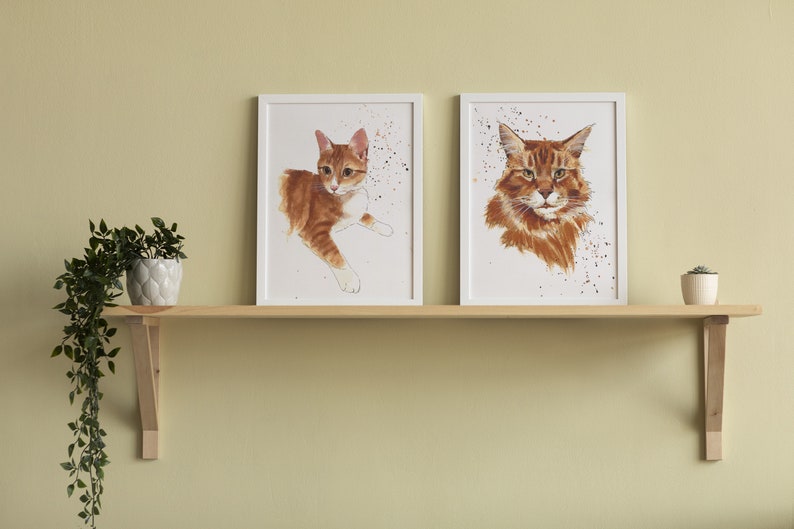 May include: Two framed watercolor paintings of orange tabby cats. The cat on the left is lying down, while the cat on the right is looking directly at the viewer. Both paintings have a white background with splattered paint.