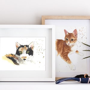 May include: Two framed watercolor paintings of cats. The painting on the left features a calico cat with green eyes looking directly at the viewer. The painting on the right features a ginger cat with white markings lying down. Both paintings have a white background with gold speckles.