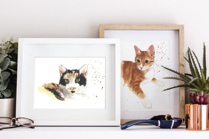 May include: Two framed watercolor paintings of cats. The painting on the left features a calico cat with green eyes looking directly at the viewer. The painting on the right features a ginger cat with white markings lying down. Both paintings have a white background with gold speckles.