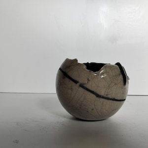 Può includere: Una piccola ciotola rotonda in ceramica con una glassa craquelé e linee nere. La ciotola ha un bordo leggermente irregolare ed è di colore beige chiaro.