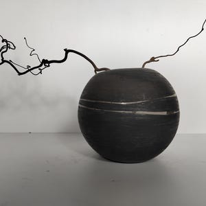 Könnte beinhalten: Eine schwarze Keramikvase mit weißen Streifen, rund und mit einer strukturierten Oberfläche. Ein getrockneter Zweig in natürlicher brauner Farbe ist in der Vase platziert.