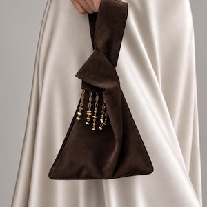 Puede incluir: Un bolso de mano de ante marrón oscuro con asa de nudo, que presenta una silueta triangular y una serie de delicadas borlas de cadena en tono dorado que cuelgan del nudo frontal. El bolso tiene una textura mate suave y un diseño sencillo.