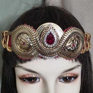 May include: A gold-colored headpiece with a central teardrop-shaped red jewel, flanked by two circular snake-themed ornaments. The headpiece is accented with red fabric and small red jewels, creating a regal and ornate design.
