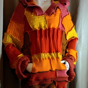 May include: A multi-colored patchwork sweater with a hood. The sweater is made of different shades of red, orange, and yellow knit fabric. The sweater has a relaxed fit and a long, flowing silhouette.