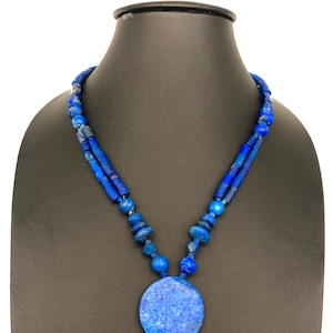 以下が含まれることがあります： 大きな丸いペンダントが付いた青いラピスラズリネックレス。ネックレスは小さな丸いビーズで作られており、銀のチェーンに通されています。